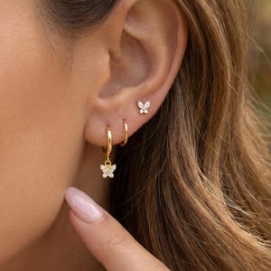 Puede incluir: Pendientes de aro y pendientes de botón dorados con diseños de mariposas. Los pendientes están adornados con pequeñas piedras brillantes. La imagen muestra los pendientes en una oreja, mostrando los delicados detalles y el diseño de la joyería.