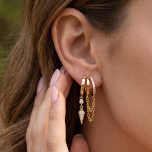 Pode incluir: Um conjunto de três brincos de argola dourados usados numa única orelha. A primeira argola apresenta um pendente com três pequenas pedras claras em forma geométrica. A segunda argola tem uma corrente dourada delicada. A terceira argola está incrustada com pequenas pedras claras.