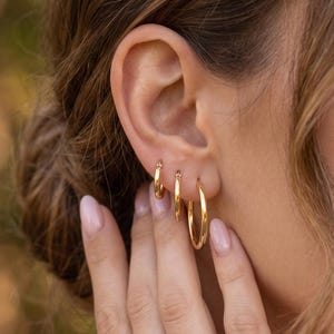 May include: Three gold hoop earrings of varying sizes worn in a single earlobe. The earrings are circular, polished, and metallic, featuring a simple continuous design. The smallest hoop is positioned at the top, followed by a medium hoop, and the largest.