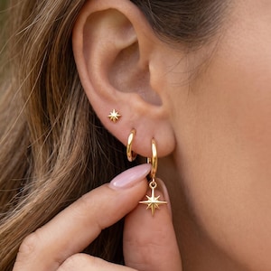 May include: A set of gold earrings, including small star stud earrings, small hoop earrings, and hoop earrings with a dangling star charm. The earrings are displayed on an ear, showcasing their design and style. The earrings are gold in color.