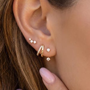 Könnte beinhalten: Nahaufnahme eines Ohrs, das mit mehreren goldenen Ohrringen geschmückt ist. Die Ohrringe umfassen kleine Diamantstecker, einen gebogenen Reifen und eine Ansammlung von drei kleinen Diamanten.