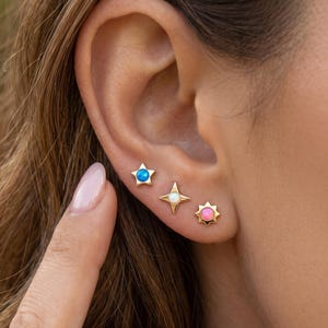 May include: Three gold stud earrings arranged in a row on an earlobe. The first is a five-pointed star with a blue center, the second is a four-pointed star with a white center, and the third is a sun-shaped stud with a pink center.