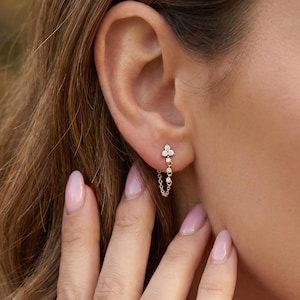 Puede incluir: Pendientes dorados con un diseño de racimo floral en el lóbulo de la oreja y una delicada cadena con detalles colgantes. Los pendientes presentan pequeñas gemas brillantes, añadiendo un toque de elegancia a la joyería.