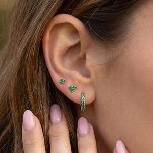Könnte beinhalten: Nahaufnahme eines Ohrs mit drei verschiedenen Arten von smaragdgrünen Ohrringen. Dazu gehören ein kleiner Stecker, ein dreieckiger Cluster und ein kleiner Creolen-Ohrring. Der Hautton ist hell, die Nägel sind hellrosa lackiert.