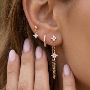May include: A set of three gold-tone earrings consisting of a small stud with a four-petaled flower charm, a thin hoop earring encrusted with small clear stones, and a stud earring featuring a flower charm connected to a chain with a square stone.
