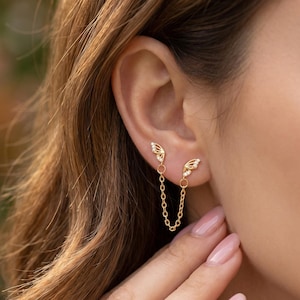 May include: A pair of gold-coloured wing-shaped earrings linked by a fine chain. The earrings are positioned on the ear, with the chain hanging down. The wings feature detailed patterns and tiny, glittering embellishments. The design is graceful and refined.