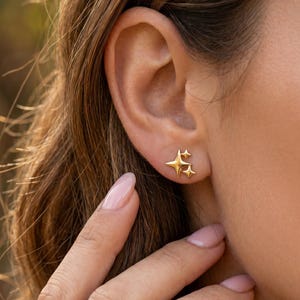 Könnte beinhalten: Nahaufnahme eines Ohrs mit goldenen sternförmigen Ohrsteckern. Die Ohrringe haben ein dreidimensionales Design mit mehreren Spitzen. Der Hintergrund ist verschwommen, was auf eine Umgebung im Freien hindeutet.