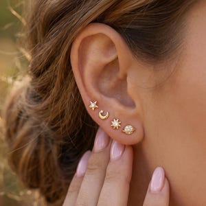 Könnte beinhalten: Goldfarbene Ohrstecker mit Himmelsmotiven: ein Stern, ein Halbmond, eine Sonne und ein Planet, jeweils mit kleinen, funkelnden Details. Die Ohrringe werden an einem Ohr getragen.
