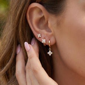 May include: Gold-toned earrings with a floral design. The set includes small stud earrings, hoop earrings, and a dangling charm earring. The earrings feature clear, sparkling stones, and are worn on the ear.
