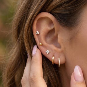 May include: A collection of five gold-toned earrings worn in a single ear. The set includes a three-stone cluster stud, a small gold ball stud, a four-stone flower-shaped stud, a plain gold hoop, and a thin gold hoop encrusted with small clear stones.
