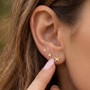 May include: Three gold-colored flower stud earrings of varying sizes are displayed on an ear. The earrings are in a graduated size, with the largest flower stud at the bottom. The earrings are a simple, elegant design.