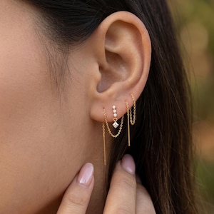 May include: A close-up shot of an ear displaying three sets of gold earrings. One set includes a diamond-shaped stud with a chain, another has three small studs, and the third has a chain and a bar. The earrings are set against a blurred background.