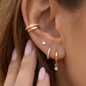 May include: A gold ear cuff featuring a row of small clear stones alongside a plain gold band, a single clear stone stud earring, a small gold hoop earring with clear stones, and a gold hoop earring with a dangling clear stone charm.