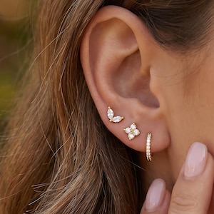 May include: A close-up of an ear displaying three gold earrings. The top earring is a small, gold butterfly shape with clear stones. The middle earring is a gold flower shape with clear stones. The bottom earring is a small gold hoop with clear stones.