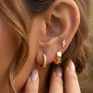 May include: A set of three gold-toned earrings consisting of one small lightning bolt stud with clear stones, one plain polished gold hoop, and one small gold hoop encrusted with a row of clear stones along the outer edge of the earring band.