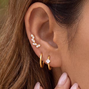 May include: A set of three gold-toned earrings consisting of one curved ear climber featuring multiple small clear stones, one plain gold hoop, and one gold hoop earring accented with a single small square-cut clear stone set in the center.