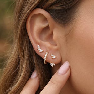 Op de afbeelding: Een verzameling gouden oorbellen met heldere edelstenen. De oorbellen omvatten een kleine hoepel, blad-vormige studs en een cluster van drie blad-vormige studs. De oorbellen worden op een oor getoond.