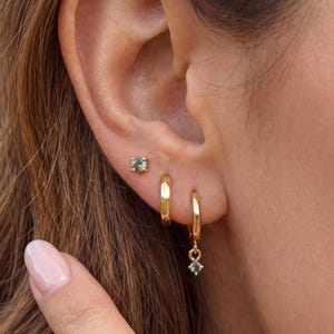 May include: A set of three gold-toned earrings consisting of one small stud with a round green gemstone, one plain gold hoop, and one gold hoop featuring a small dangling round green gemstone, all worn in a single earlobe arrangement.