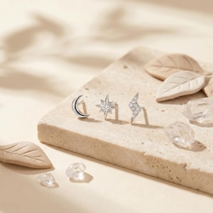 May include: Three silver-toned stud earrings featuring a crescent moon, a multi-pointed star with a central clear stone, and a lightning bolt shape encrusted with small clear stones. The earrings are arranged in a row on a textured surface.