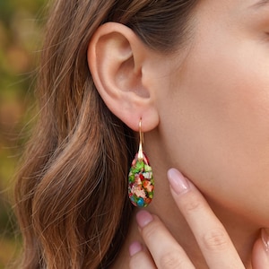 May include: A teardrop-shaped earring featuring a multicoloured stone with vibrant inclusions of green, red, blue, and white. The stone is suspended from a thin, polished gold-toned metal hook that passes through the earlobe, creating a dangling effect.