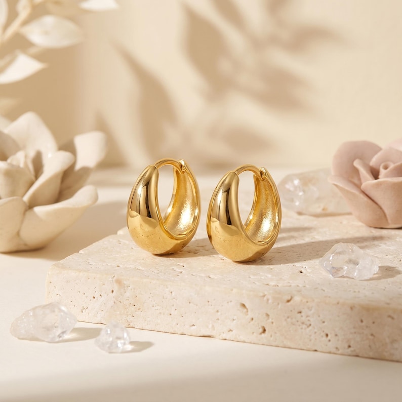 May include: A pair of polished gold hoop earrings with a thick, rounded, and smooth surface. The earrings feature a hinged closure mechanism and a high-shine metallic finish that reflects light, creating a bright and reflective appearance.
