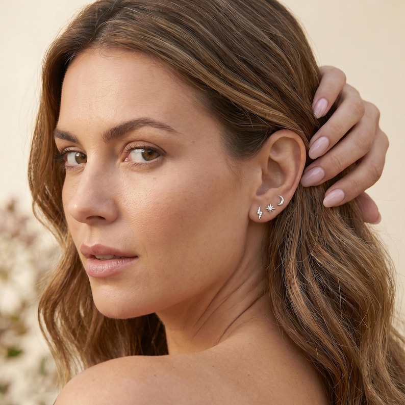 May include: A set of three small silver-toned stud earrings worn in one ear. The earrings feature a lightning bolt shape with embedded stones, a four-pointed star with a central stone, and a simple crescent moon shape, all in a metallic finish.