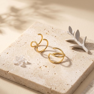 Pu&ograve; includere: Due orecchini dorati dal design minimalista sono esposti su una superficie chiara e strutturata. Uno presenta un design aperto e a spirale, mentre l'altro ha un aspetto curvo e stratificato. Sono presenti anche un piccolo cristallo trasparente e un ramo decorativo bianco.