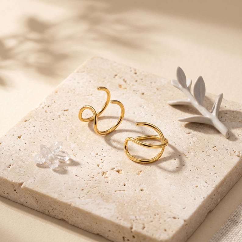 Pu&ograve; includere: Due orecchini dorati dal design minimalista sono esposti su una superficie chiara e strutturata. Uno presenta un design aperto e a spirale, mentre l'altro ha un aspetto curvo e stratificato. Sono presenti anche un piccolo cristallo trasparente e un ramo decorativo bianco.