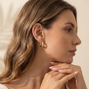 May include: A small, polished gold hoop earring with a thick, rounded profile. The earring features a smooth, reflective surface and a circular shape, securely fastened through the earlobe, showcasing a simple and classic jewelry design.