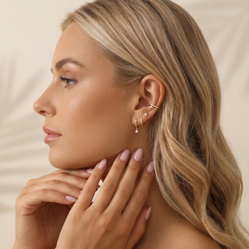 Pode incluir: Um conjunto de brincos dourados composto por um ear cuff de banda dupla no topo, um pequeno brinco de pino no meio e uma argola delicada com uma &uacute;nica pedra transparente pendente na parte inferior, usados num &uacute;nico l&oacute;bulo.
