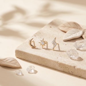 May include: Three gold stud earrings featuring a crescent moon, a star with a central clear stone, and a lightning bolt shape encrusted with small clear stones. The earrings are arranged in a row on a textured, light-colored stone surface.