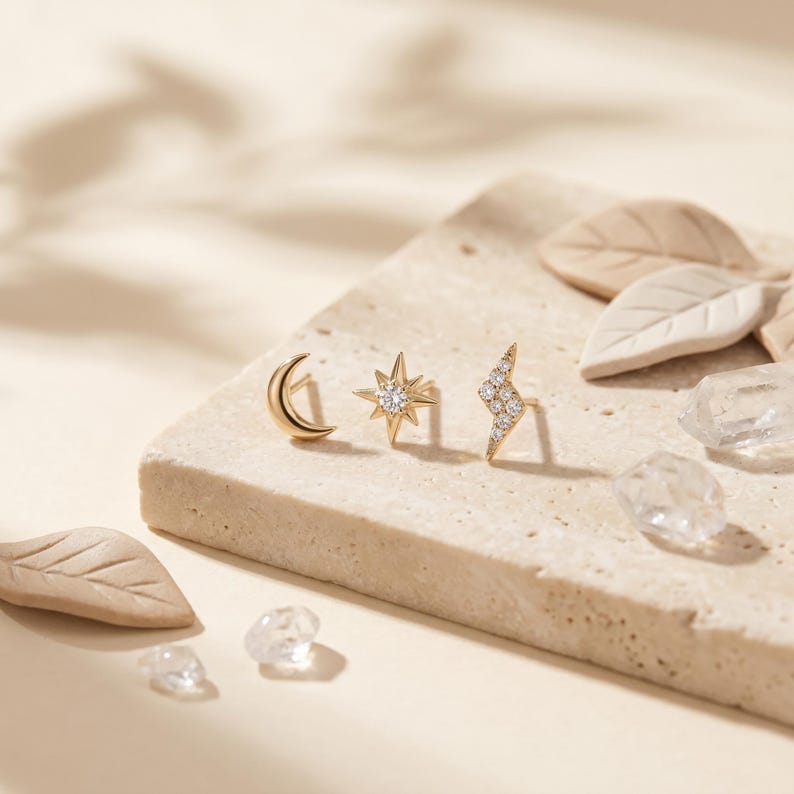 May include: Three gold stud earrings featuring a crescent moon, a star with a central clear stone, and a lightning bolt shape encrusted with small clear stones. The earrings are arranged in a row on a textured, light-colored stone surface.