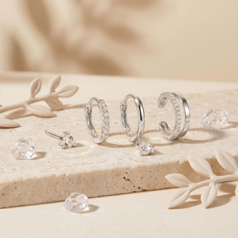 Pode incluir: Um conjunto de joias em tom prateado, incluindo um pequeno brinco de pino com uma pedra clara, dois pequenos brincos de argola com pedras claras e um ear cuff de banda dupla com uma fila de pedras claras, tudo com acabamento polido.