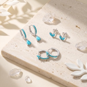 May include: A collection of silver-toned jewellery featuring bright turquoise stones. The set includes a pair of hoop earrings with teardrop pendants, a pair of small leaf-shaped stud earrings, and a pair of hoop earrings with a three-stone cluster.
