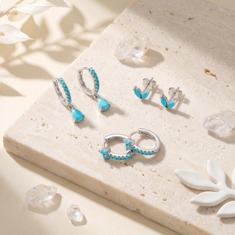 May include: A collection of silver-toned jewellery featuring bright turquoise stones. The set includes a pair of hoop earrings with teardrop pendants, a pair of small leaf-shaped stud earrings, and a pair of hoop earrings with a three-stone cluster.