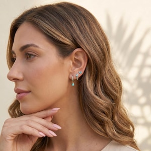 May include: A set of three gold-toned earrings featuring small turquoise-coloured stones. The set includes a small hoop earring with a dangling teardrop charm, a second small hoop with a cluster of stones, and a tiny stud earring with a butterfly shape.