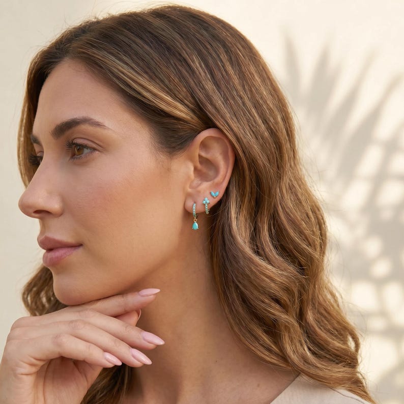 May include: A set of three gold-toned earrings featuring small turquoise-coloured stones. The set includes a small hoop earring with a dangling teardrop charm, a second small hoop with a cluster of stones, and a tiny stud earring with a butterfly shape.