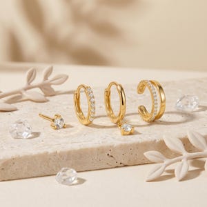 Pode incluir: Um conjunto de joias em tom dourado, incluindo um brinco de pino com uma pedra clara, dois pequenos brincos de argola com pedras claras e um ear cuff de banda dupla com pedras claras. As pe&ccedil;as apresentam metal polido e detalhes brilhantes.