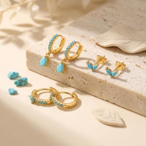 May include: A set of gold-toned earrings featuring turquoise accents. The collection includes two hoop earrings with teardrop charms, two small hoop earrings with embedded turquoise stones, and two stud earrings shaped like small leaves with turquoise.