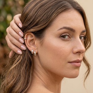 Puede incluir: Primer plano de una oreja adornada con joyas de plata. Se observa un peque&ntilde;o pu&ntilde;o de oreja y dos pendientes de aro plateados. La mano de la persona toca su cabello. El fondo est&aacute; borroso, posiblemente una escena al aire libre.