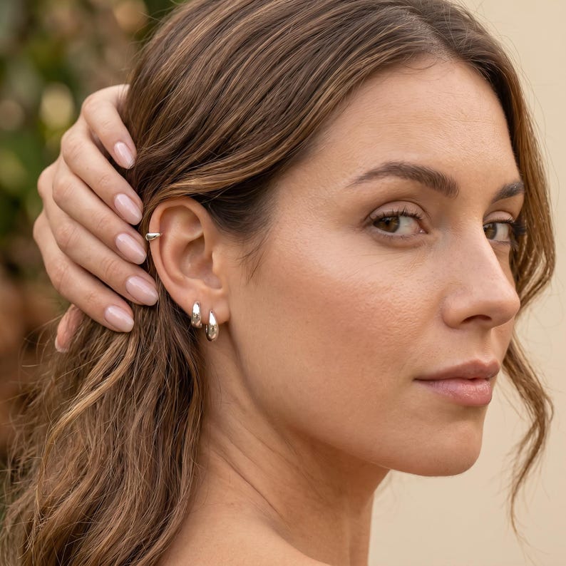 Puede incluir: Primer plano de una oreja adornada con joyas de plata. Se observa un peque&ntilde;o pu&ntilde;o de oreja y dos pendientes de aro plateados. La mano de la persona toca su cabello. El fondo est&aacute; borroso, posiblemente una escena al aire libre.