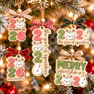 Puede incluir: Cinco adornos navideños de madera con un personaje de bola de masa con astas. Cada adorno muestra el año 2026 en rosa, verde y blanco, con textos que incluyen My Squishy Era, The Year of Squishies y Merry Squishmas.
