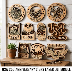 May include: A collection of laser-cut wooden signs with patriotic themes. Designs include eagles, stars, and text such as "Celebrating 250 Years" and "USA 1776-2026". The signs are displayed on a wooden surface.