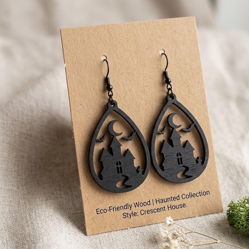 Puede incluir: Un par de pendientes colgantes de madera negra con un dise&ntilde;o recortado de una casa encantada, una luna creciente y murci&eacute;lagos. Los pendientes est&aacute;n sujetos a una tarjeta beige con el texto Eco-Friendly Wood | Haunted Collection Style: Crescent House.