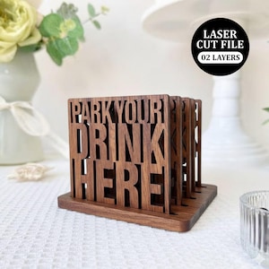 Puede incluir: Posavasos de madera marrón oscuro con las palabras "PARK YOUR DRINK HERE" recortadas. Los posavasos están en un soporte a juego. La imagen también incluye un círculo negro con el texto "LASER CUT FILE 02 LAYERS".