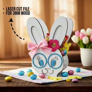 Może przedstawiać: Biały drewniany koszyk wielkanocny w kształcie królika z niebieskimi okularami, różową kokardą i różowym kwiatem. Koszyk jest wypełniony wielkanocnymi smakołykami i otoczony czekoladowymi jajkami i cukierkami. Tekst brzmi: "LASER CUT FILE FOR 3MM WOOD."