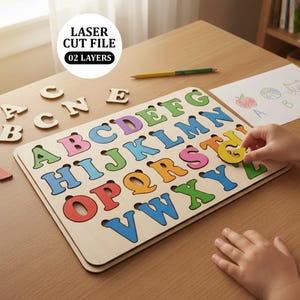 May include: Wooden alphabet puzzle with colorful letters. The puzzle board features the letters A through Z in various colors. A child's hand is placing the letter "G" into the puzzle. The image also includes the text "LASER CUT FILE 02 LAYERS".