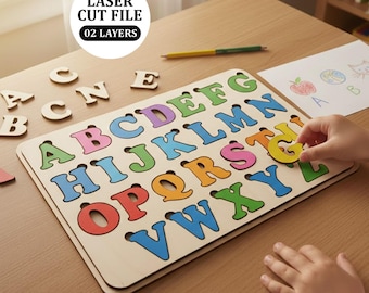 May include: Wooden alphabet puzzle with colorful letters. The puzzle board features the letters A through Z in various colors. A child's hand is placing the letter "G" into the puzzle. The image also includes the text "LASER CUT FILE 02 LAYERS".