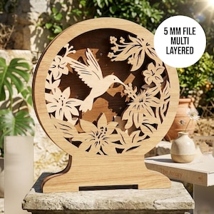 May include: A wooden, multi-layered, round decorative piece featuring a hummingbird and floral design. The hummingbird is in flight, surrounded by intricately cut flowers and leaves. The text "5 MM FILE MULTI LAYERED" is in a white circle on the right side.