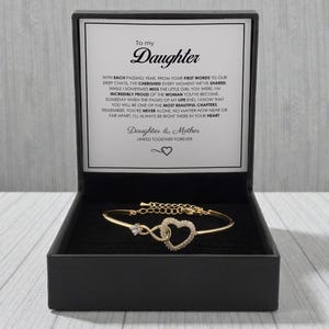 Puede incluir: Una pulsera de tono dorado con un dije en forma de corazón incrustado con pequeñas piedras transparentes, presentada en una caja de regalo negra. La tapa interior muestra un mensaje impreso dirigido a una hija, expresando sentimientos sobre su vínculo.